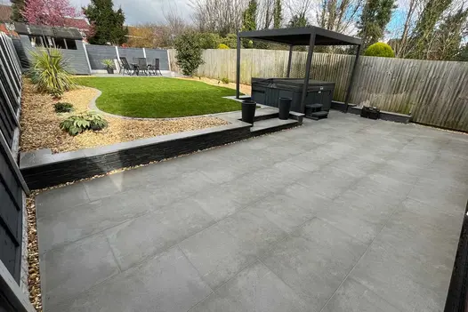 Back garden with large oblong patio paved with slabs, hot tub under Proteus grey aluminium pergola, and lawn surrounded by gravel.