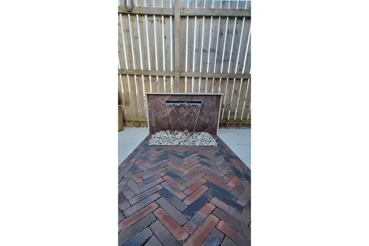 In front of fence, water cascades into gravel from wide spout in low wall faced with Abbey Dark Multi clay pavers. 