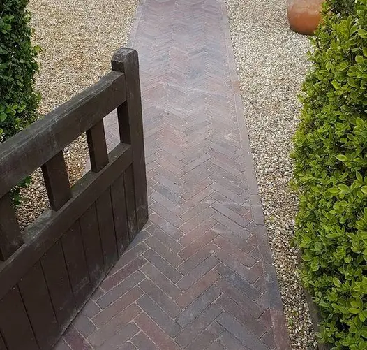 Wooden gate set into hedge opens over straight path of Abbey Dark Multi clay pavers laid herringbone pattern between gravel areas. 