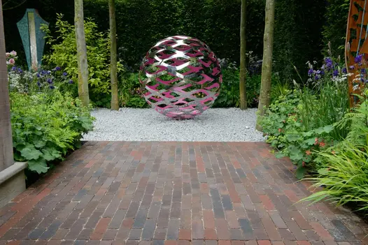 Pink and silver perforated sphere sits on white gravel, approached by wide path of Abbey Dark Multi clay bricks between beds.