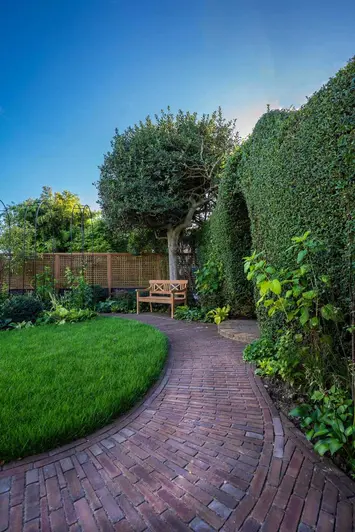 Curved path of Abbey Dark Multi Brick Pavers runs between clipped hedge and lush lawn, past a wooden bench in fenced garden.