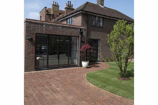 Wide driveway of Abbey Dark Multi clay pavers laid herringbone curves around grass and narrows on approach to side door of house.