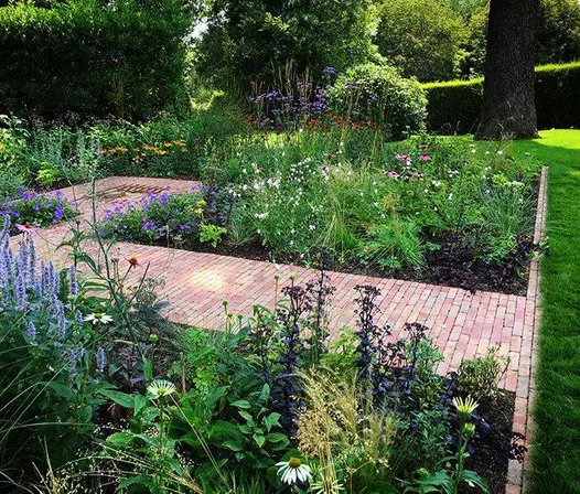 Flower beds around Seville clay paving path in rectangular area by hedge in large lawn with mature trees.