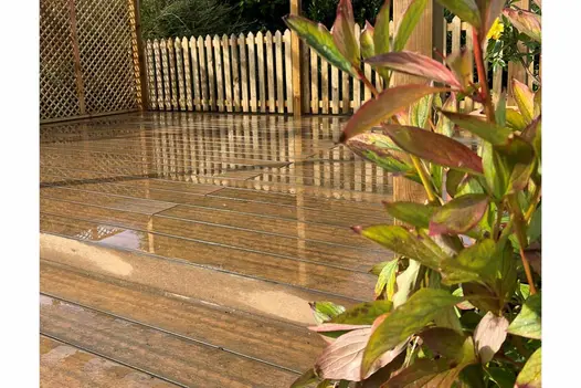 Low view of wet garden decking, warm teak brushed composite shows colour and texture, red and green plants in foreground.
