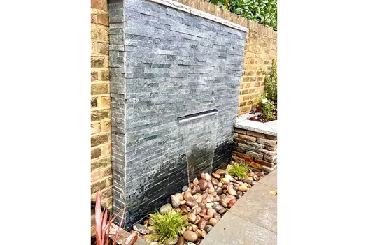 Brick wall interrupted by proud section of Green Slate stone cladding with letterbox spout emptying water onto pebbles below.
