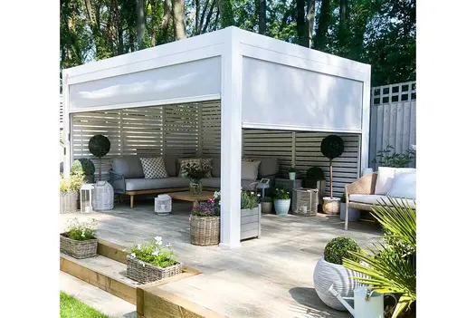 Proteus White Aluminium pergola with optional sides stands over outdoor furniture and planted pots on raised patio. 
