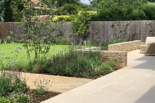 Florence beige porcelain patio, partially edged with dry stone walls, accessed by clay paver path between flower beds and lawn.