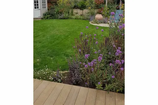 Amber composite decking boards laid perpendicular to curve-edged lawn and flower bed. Round patio with garden chairs in background.