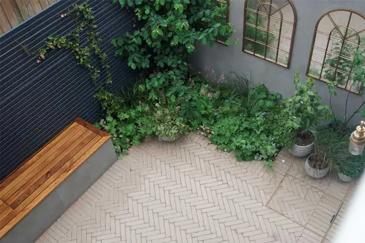 View from above of small courtyard with 3 arched mirrors on blue wall above foliage-filled bed, paved with stone grey clay bricks.