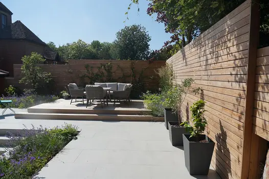Raised decking area in the corner of a country garden with timber privacy screens on two sides and surrounded by a large porcelain patio.