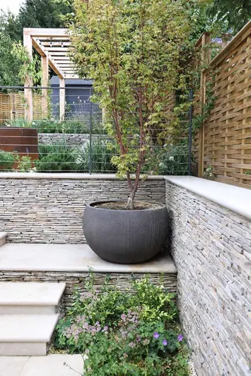 View from the corner of a garden, a large plant pot sits on Jura Beige bullnose steps, clad with cobblestones and surrounded by plants.