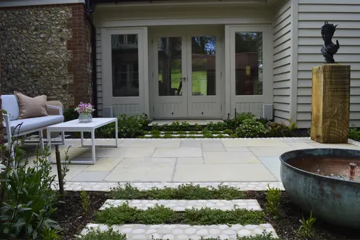 Outdoor furniture on oblong patio of Antique Yellow limestone paving edged with cobbles outside taupe-coloured french doors.
