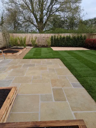 Fenced garden with dog-leg lawn on 2 sides of large paved area of Antique Yellow limestone with L-shaped sleeper-built beds.