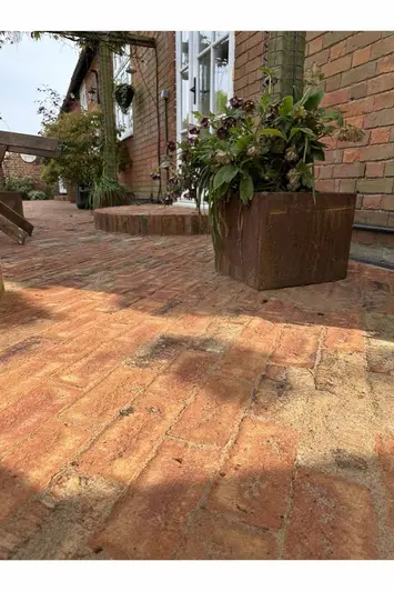 Low angled camera shot of cotswold blend belgian bricks shows red brick house in the background after a steel corten plant pot.