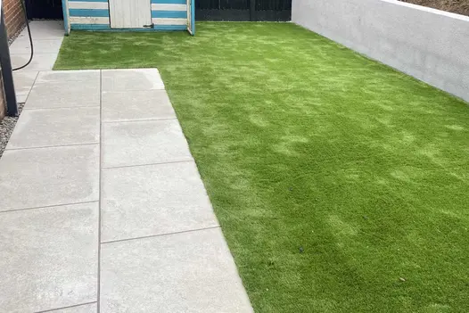 Astor Grey porcelain paving set into artificial lawn leads to path by wall to striped garden shed. Built by Coastal Gardens.