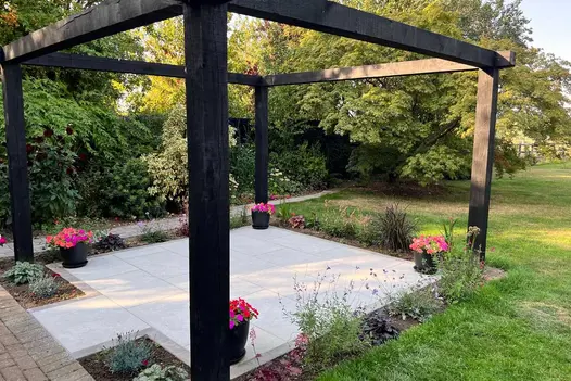 Pergola sits on top of small area paved with Astor Grey Porcelain Paving with potted pink plants and flower beds surrounding.
