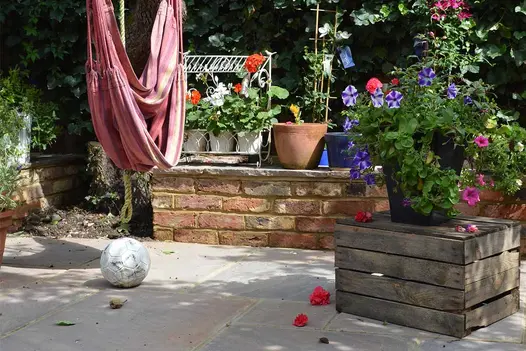 Football and fallen blooms on Autumn Brown Indian sandstone slabs laid in leafy corner of fenced garden, with pots on raised ledge.