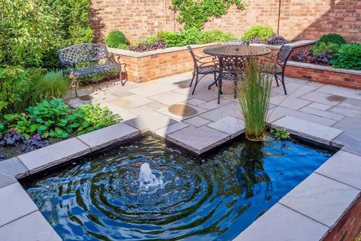 Bubbling L shaped pond sits raised in the middle of autumn brown patio, topped with matching indian rock-faced coping stones.