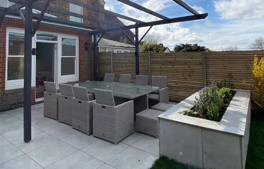 Patio doors leading out to a porcelain patio dining area set underneath a pergola with a raised planter bed and slatted fencing panels.