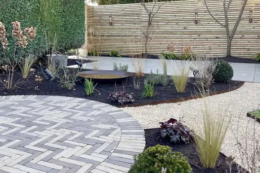 Round paved area of Gromo Antica brick paving abuts curved planted beds and gravel path to rectilinear paved area next to fence.
