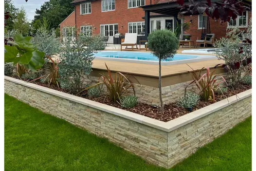 Brick wall capped with Beige sawn sandstone coping stones retains planted bed on 2 sides of raised patio with swimming pool.