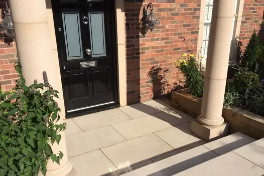 two steps down to front door utilising the beige smooth sandstone paving slabs with two large pillars either side of the door.