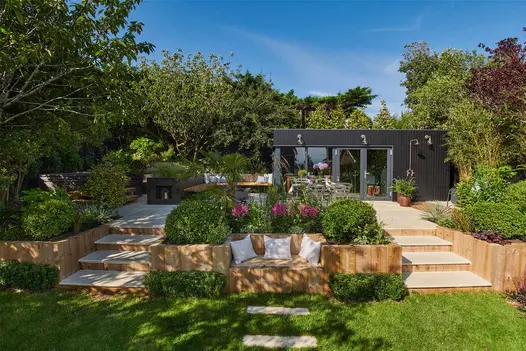 2 sets of Beige Sawn Sandstone steps rise either side of raised bed to matching patio with garden room amidst lush planting.