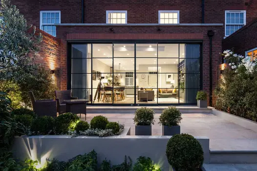 Lit open-plan interior looks out over patio of Beige sandstone smooth paving slabs with potted topiary and wall shrubs.