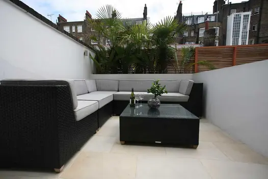 Dark modular furniture sits on Beige smooth sandstone patio enclosed by low white wall, with slatted fence behind. Built by HCL.