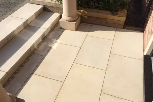 close view of 2 steps of beige smooth sandstone stepping down onto small patio for front door area. Pillars either side.