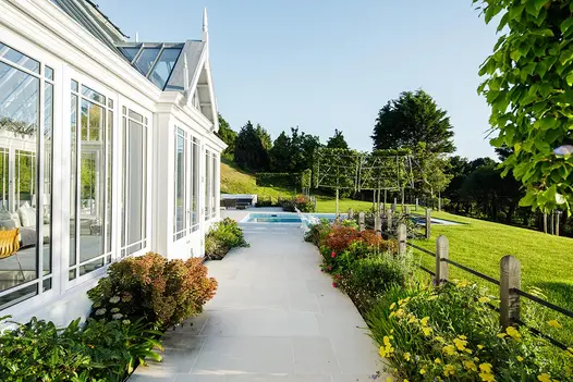 Beige smooth paving slabs run between side of gabled conservatory and lawn with planted border towards swimming pool.