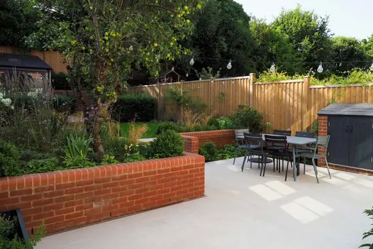 Fenced garden with low brick wall forming boundary of L-shaped patio in Beige smooth sandstone paving. Lawn and planting behind.