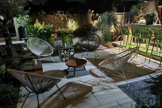 Garden paved with Beige smooth sandstone slabs and clay pavers, cluttered with seats, fire pit, log store, bar and potted palms.