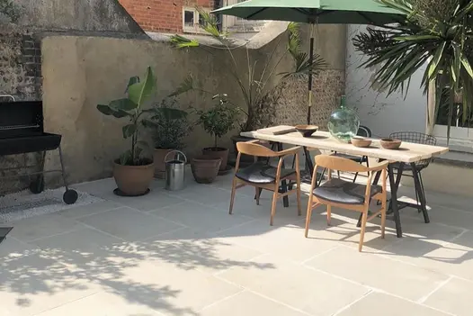 Dining set with parasol sits on Beige smooth sandstone paving with potted plants. Brick and cement rendered wall behind.