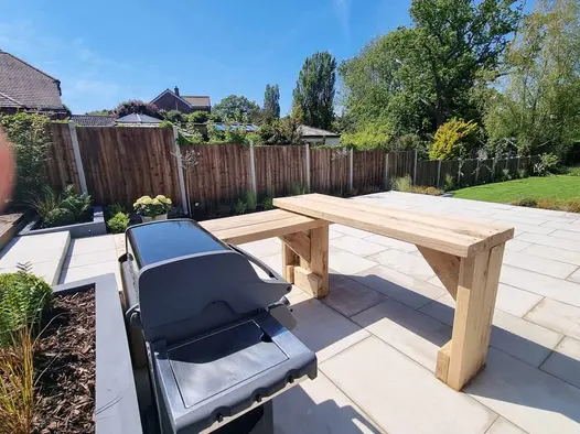 Gas barbecue and intersecting wooden tables sit on patio of Beige smooth sandstone paving in fenced garden with lawn beyond.