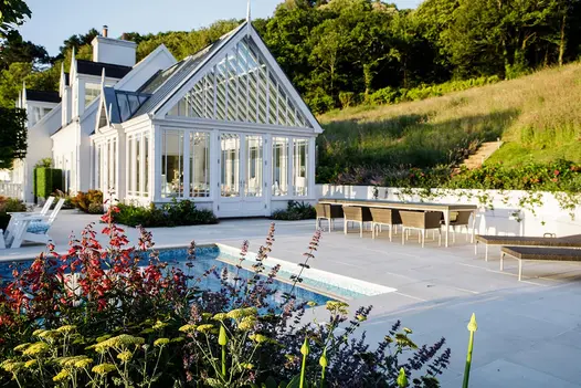 Grass slopes down behind large area of Beige smooth sandstone patio paving between white-painted conservatory and swimming pool.