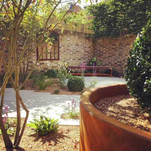 Corner of high-walled garden with Beige smooth sandstone plank paving and gravel-mulched beds with scattered planting.