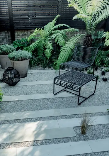 Metal chair and footstool sit on paved area of gravel with page smooth sandstone plank paving. Tree ferns next to boundary wall.