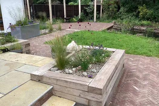 Rectangular raised bed of sleepers on edge of raised patio with path of Bexhill brick pavers turning 90 degrees at its corner. 