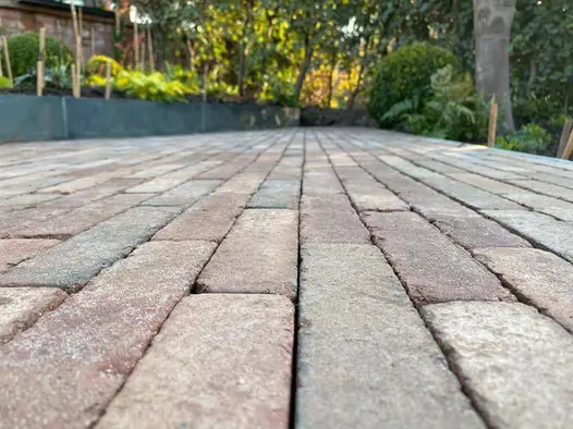 Ground level view of Bexhill brick paving leading towards blurred trees in distance, next to raised beds and mature planting.