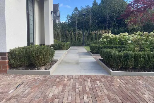 In modern garden design Bexhill Clay Paving abuts cream-coloured path between stone-edged flower beds in front of house on left.