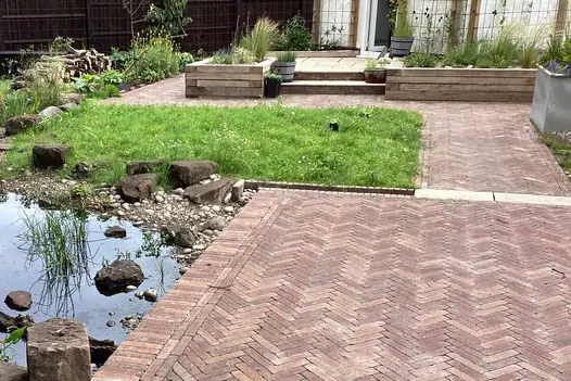 Patio of Bexhill clay pavers, laid herringbone with soldier course edge, next to natural pond with pebbly approach from small lawn.