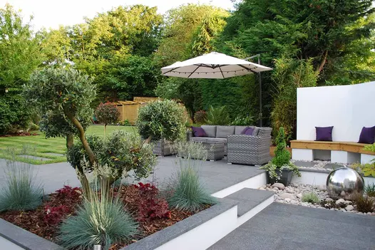 L-shaped patio of Black granite paving with cloud-pruned tree in raised bed opposite rattan sofa and free-standing parasol.