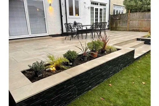2 raised beds, separated by steps down to lawn, form edge of cream-coloured patio. Their walls are faced with Black slate cladding.