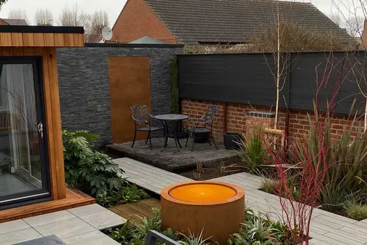 Black slate clad wall with inset Corten steel panel forms backdrop to patio in garden with studio, decking paths and water feature.
