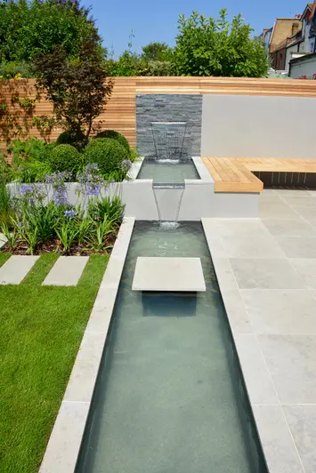 Waterfall feature with black slate cladding and porcelain, cascading water across two levels into a pond that divides the lawn and patio.