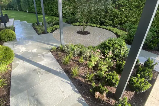 Blue Kota Limestone paving runs between planted beds towards large hedged lawn, past circle of clay pavers with central shrub.