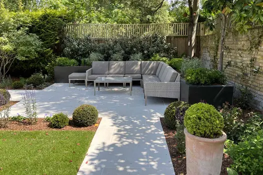 L-shaped outdoor sofa sits with back to raised bed on Florence White porcelain paving square in corner of walled and fenced garden.