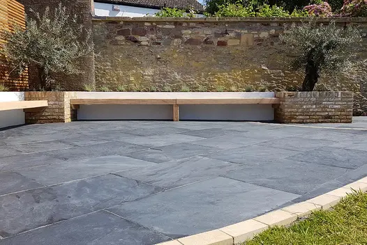 Curved Brazilian black slate patio edged with buff bricks in corner of walled garden with raised beds and inbuilt benches.