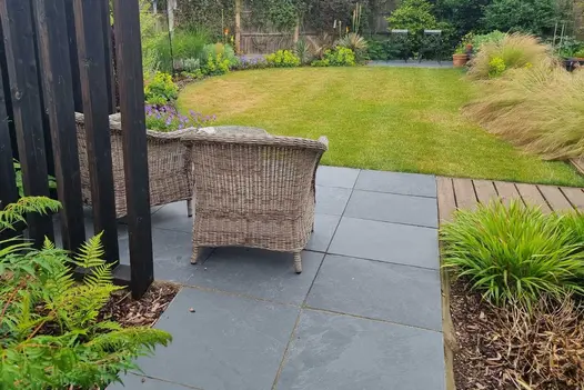 Rattan chairs sit on 600x600mm Brazilian black slate paving next to curved lawn with planted borders. Design by Paul Slater.